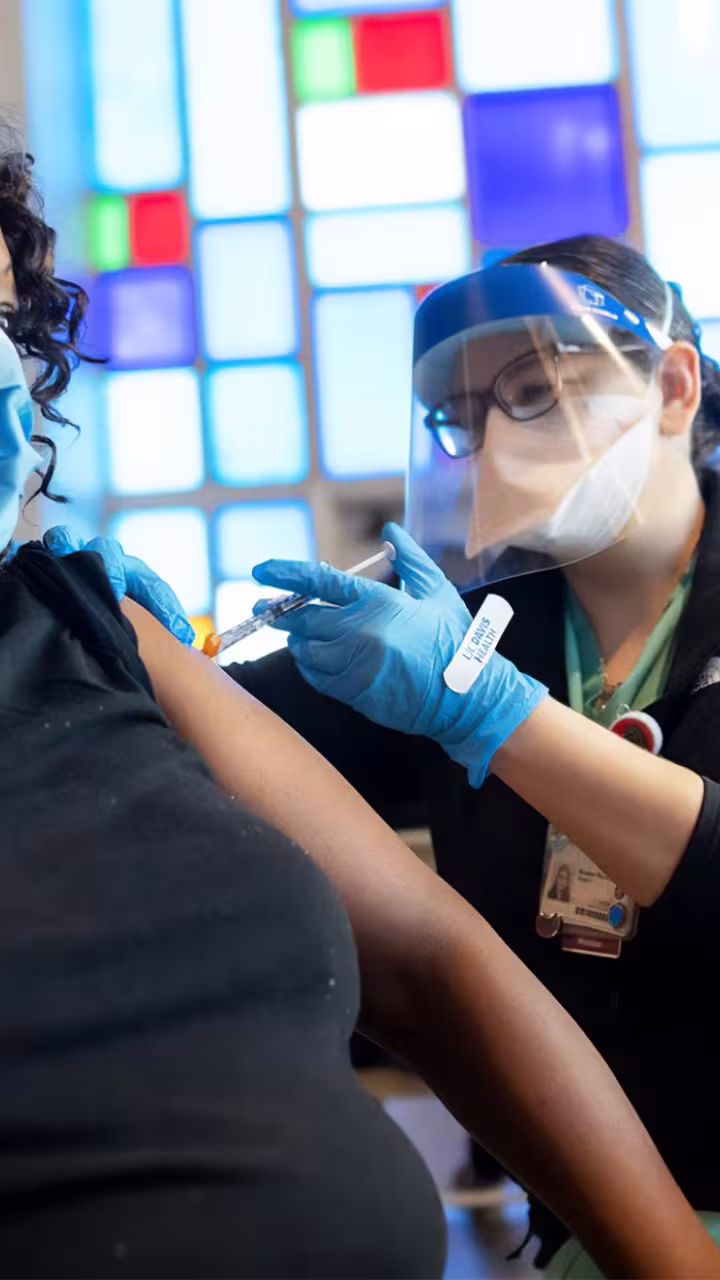 A healthcare worker in protective gear administers a COVID-19 vaccine to a masked individual wearing a black shirt, with a stained-glass window in the background.