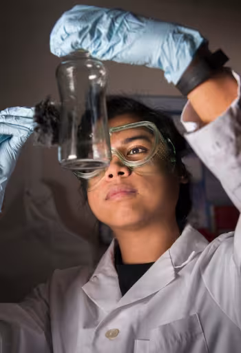 A student examines chemicals through a beaker while wearing a lab coat and goggles. 