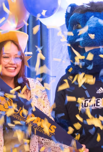 Two prospective UC Davis students pose with Gunrock the Blue Horse Mascot of the university while confetti is thrown in the air.