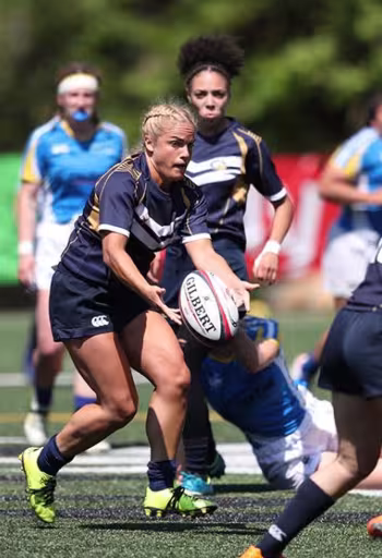 Woman in navy rugby kit passing ball as opponent dives to tackle on grassy field