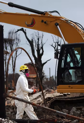 Two workers in white hazmat suits beside a yellow excavator at a muddy, bare-treed site