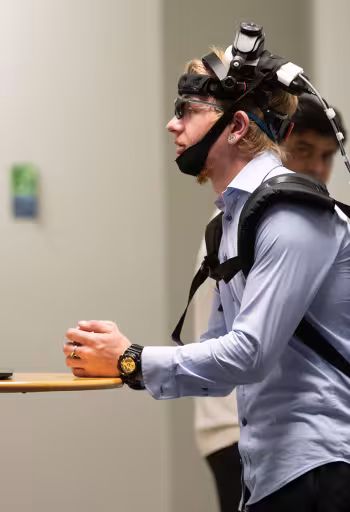 Person wearing head-mounted tracking gear and backpack, leaning on a table in a hallway.