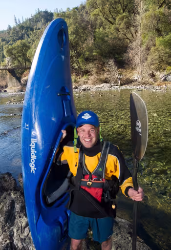 Smiling man in life jacket holding paddle beside vertical blue kayak on riverbank