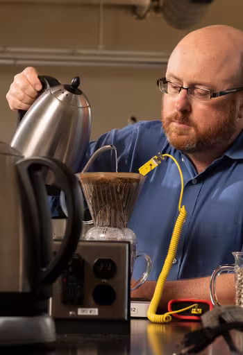 A bearded man in a blue button-down shirt and glasses concentrates while pouring hot water from a stainless-steel kettle over coffee grounds in a V60 plastic coffee dripper. He is surrounded by numerous scientific instruments and coffee tools.  