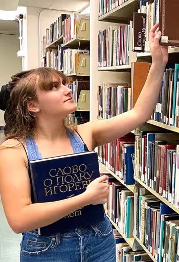 A shelf in Peter J. Shields Library displaying books with titles printed in Cyrillic. UC Davis Russian minor Ryleigh Praker reaches for a book while holding a blue book with a Russian title.