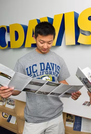 Student in UC Davis T-shirt reading a brochure under large blue-and-gold UC Davis letters