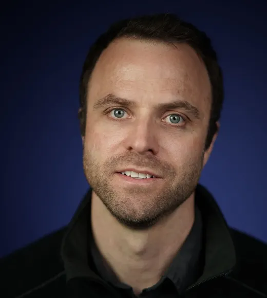 Smiling man with short dark hair and a beard, against a dark blue background.
