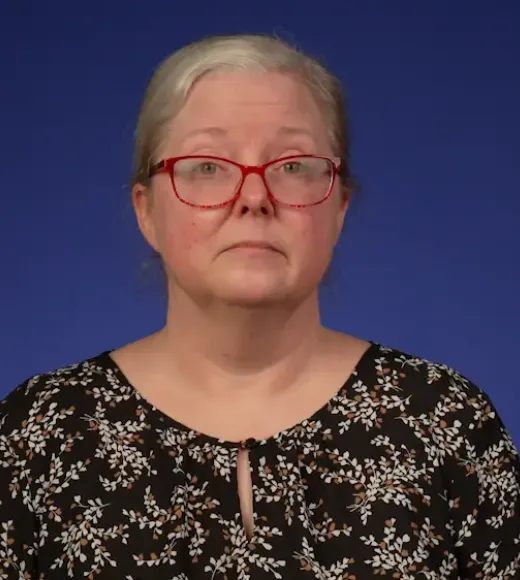 Headshot portrait of a person gray hair, red glasses, floral blouse against blue background