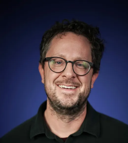 Smiling person in glasses, scruffy beard, black shirt — studio headshot, blue gradient background