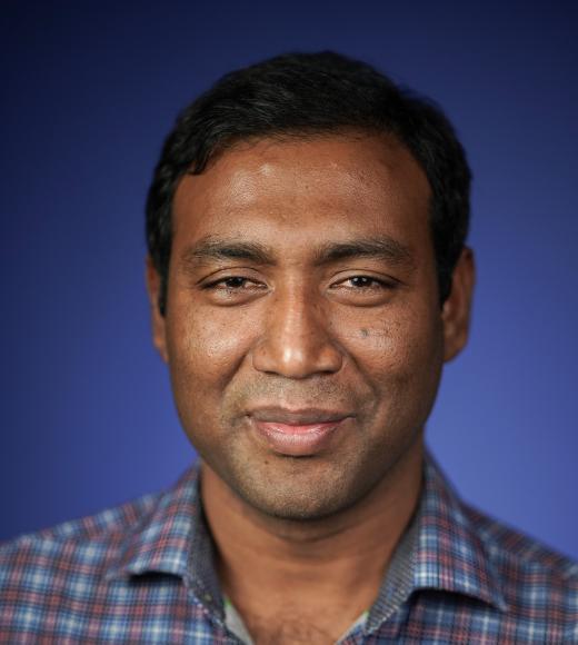 Smiling man with dark hair in a checked shirt against a blue background.