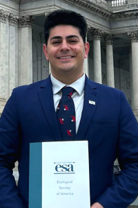 Man in a navy suit with dark hair smiles in front of a government building