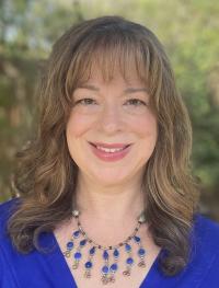 Headshot of a woman wearing a blue shirt and necklace, smiling at the camera. She is standing in front of green foliage.