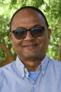 Smiling middle-aged man in sunglasses and light-blue shirt outdoors with green foliage, photo