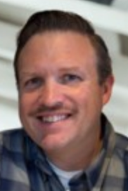 Smiling man with a mustache wearing a plaid shirt, in a bright indoor setting.