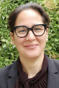 Smiling person with black glasses and dark blazer, outdoor photo with green foliage background