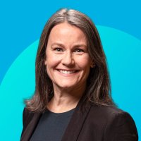 Headshot of smiling woman with long brown hair, black blazer, teal background.