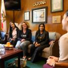 Four students sit on a couch in an office with flowers and a bowl of oranges while a politician listens.