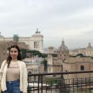 Katharine wears a cardigan and stands in front of a railing and a city scape with domes and buildings of various heights. 