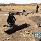 A student wearing a sunhat kneels in front of a trench on a sandy landscape. They are surrounded by tools, a team, and mountains