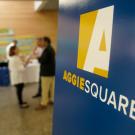 "Aggie Square" logo sign in foreground of meeting.