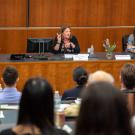 Woman gestures with pointed finger in the air, at center of dais.