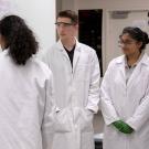 Students in lab coats stand in front of a white board.