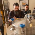 Four men watch another man working with a stainless steel vat