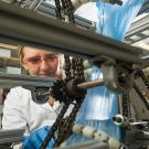 Woman in lab coat and goggles adjusts tabletop machine made of gears, chains and metal bars