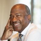 Chancellor Gary S. May at desk, smiling, hand to cheek, no jacket.