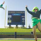 Photo: A woman and a man dressed in several pieces of green clothing pose in front of the Aggie Stadium scoreboard