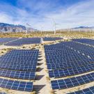 large area in desert covered by solar panels with windmills and mountains in background