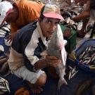 Man holding a big fish to the camera in a fish market with baskets of fish in the background