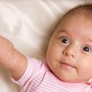 Infant girl with brown hair and brown eyes lies on her back,  reaching out one arm.