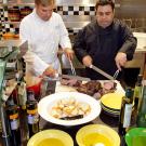 Two men cooking, with bread and olive oil in the foreground