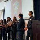 Students stand in a line and clap while looking at a projector screen.