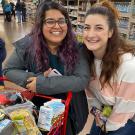 Two women stand next to full grocery cart.