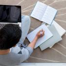 A student sits on the floor with a laptop and notebooks.