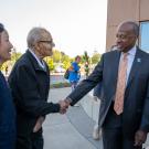 Elderly man with cane shakes hands, smiling with a suited official outdoors