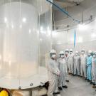 A large translucent tank fills the left side of the photo. A group of people in coveralls, helmets and facemasks stands against the wall. 