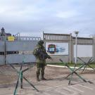 Four armed men in green uniforms, helmets and goggles stand in front of a concrete fence. 