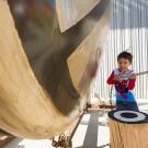 Child in superhero costume hitting a large gong sculpture 