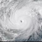 Dense white cloud seen from space spirals outwards from the center of the image. A coastline is barely visible through the cloud. 