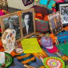 Framed photos and sculptures of sugar skulls sit atop an altar with a multicolor table cloth in an indoor setting.