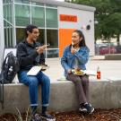 Two students sit outside Latitude Dining Commons eating food with chopsticks. They are facing the camera and angled towards each other on a sunny day. 
