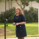 A blonde woman in a blue dress outdoors. To the left is the back of a video monitor and on the right is a blue sign slightly out of focus. 