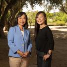 Two women photographed outdoors. The woman on left is wearing a blue shirt and facing the camera, a younger woman to the right is side on with her head turned towards the camera. 