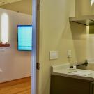 Through a kitchen doorway, Keith Graeber stands talking in living room with unseen person. A big screen tv glows blue in background