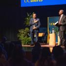 An audience inside a theater gives a standing ovation to a man on the theater stage, his hand over his heart in appreciation. Standing next to them is Chancellor Gary S May. 