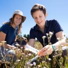 Two women in dark blue shirts kneeling in a meadow. One is holding a plastic tube while the other looks on. 
