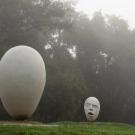 Two white abstract sculptures—one egg-shaped, one carved head—on a grassy mound in fog